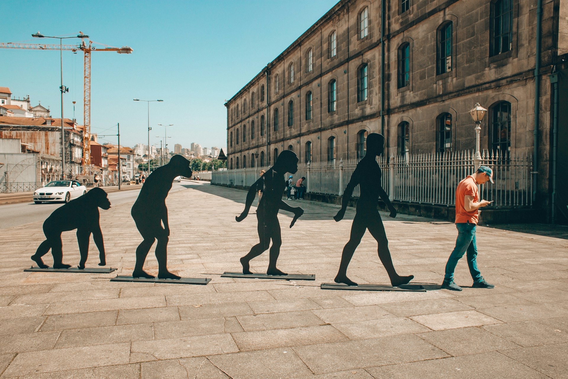 An outdoor art display of silhouettes of the progression from ape to human, with an actual person standing at the end of the progression, looking down at their phone.